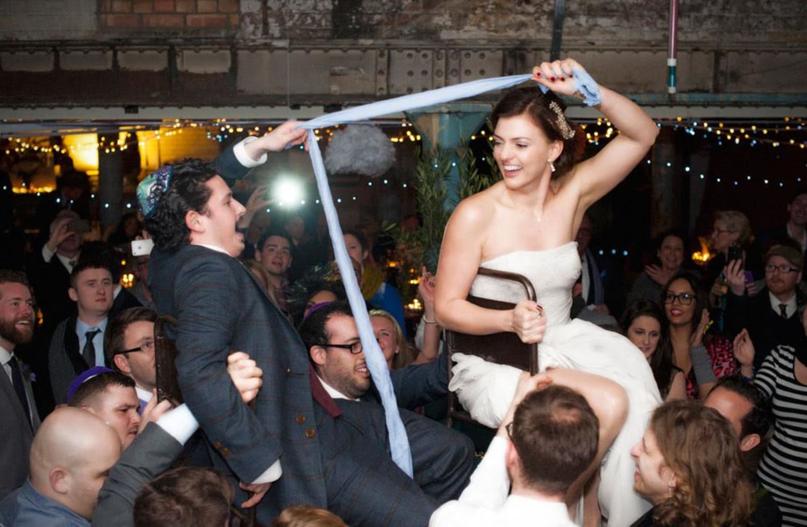 bridebook.co.uk-smashing-the-glass couple being lifted up on a chair holding a blue cloth between them
